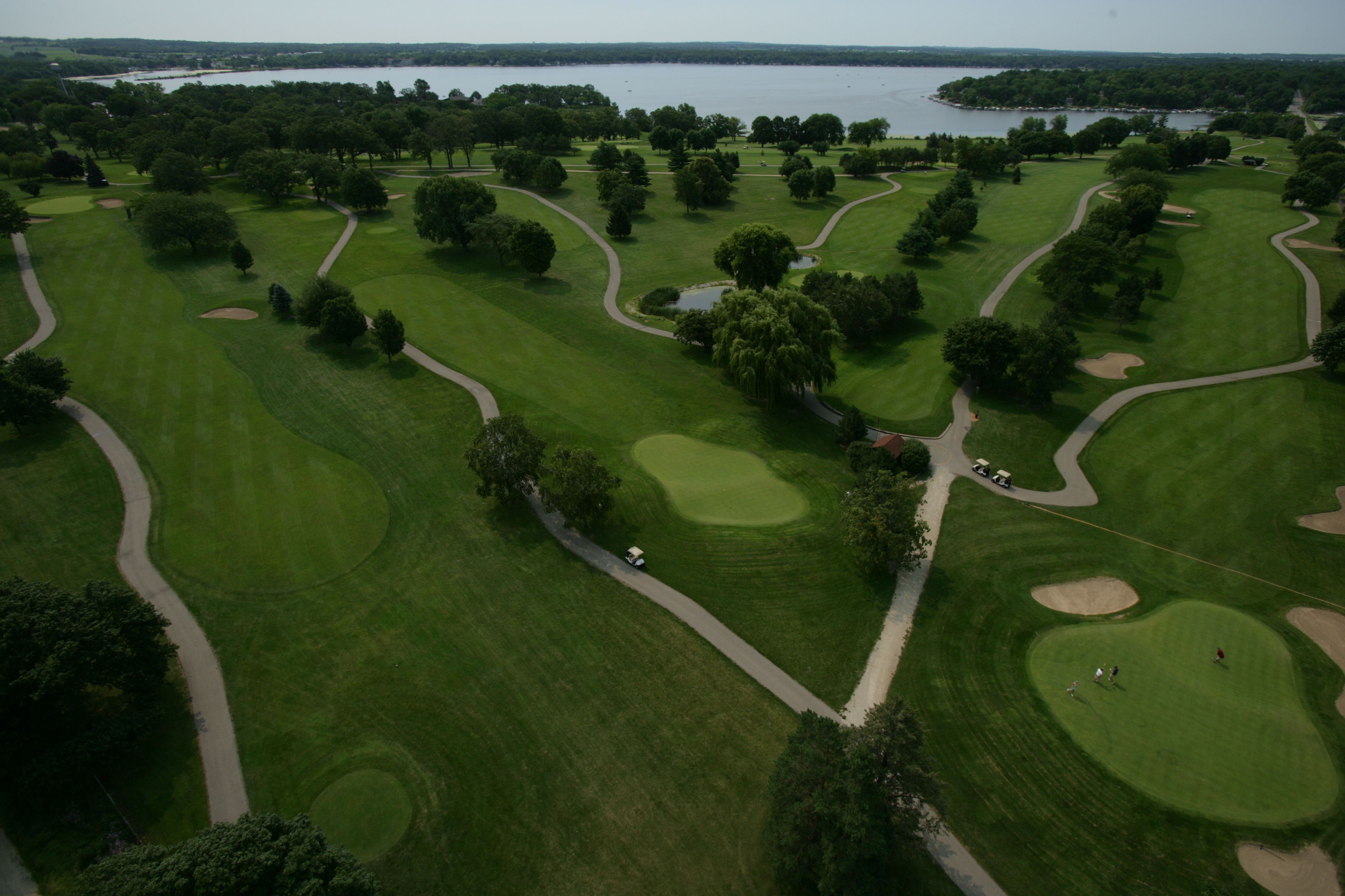 Vue aérienne d’un parcours de golf avec plusieurs greens, des bunkers de sable et une étendue d’eau en arrière-plan.