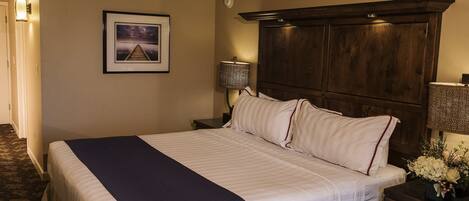 Quarto de hotel com uma cama grande, cabeceira de madeira, mesa de cabeceira com abajur, quadro na parede e arranjo floral na mesa de cabeceira.