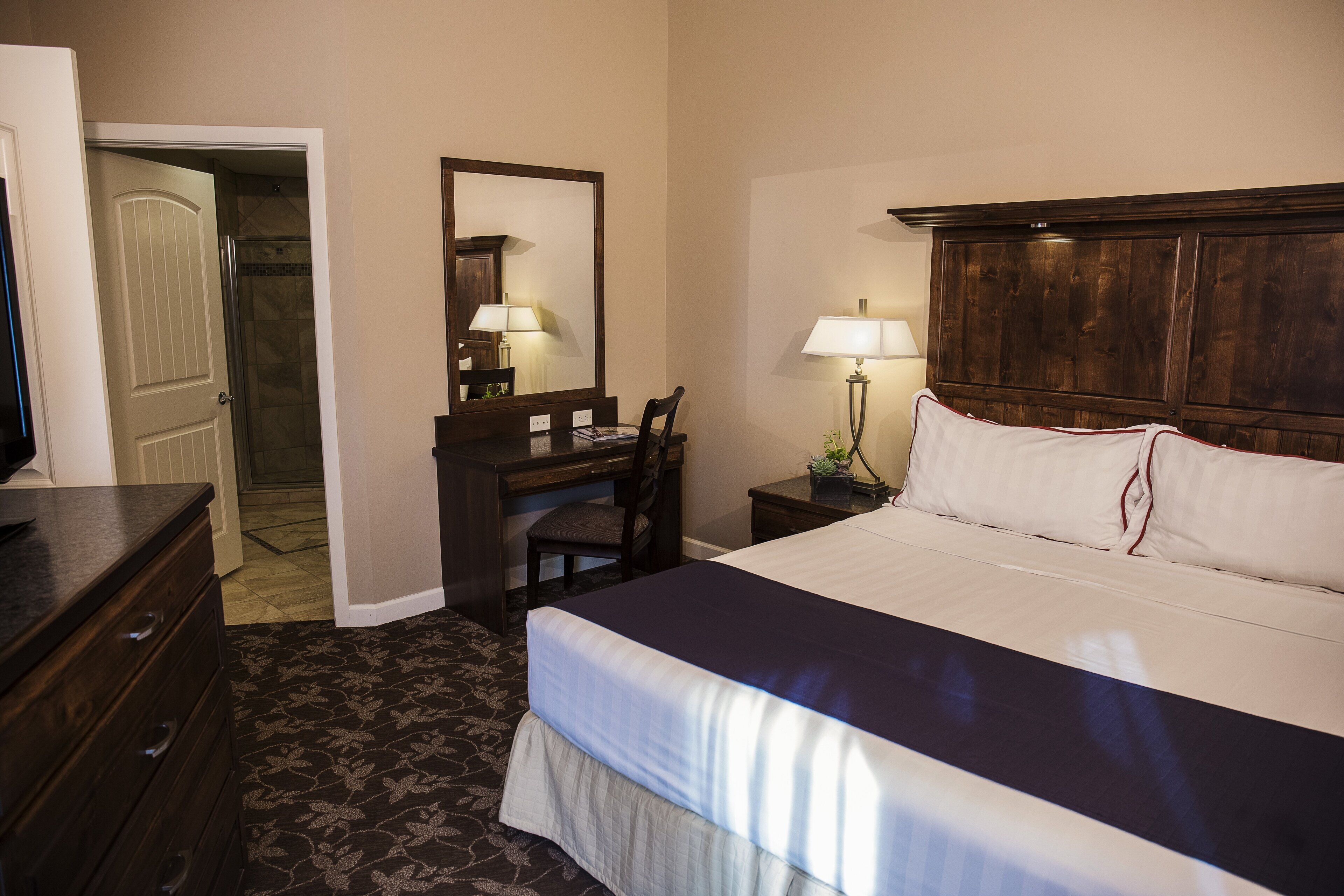 Habitación de hotel con cama, escritorio, silla, cómoda, espejo y mesita de noche con lámpara.