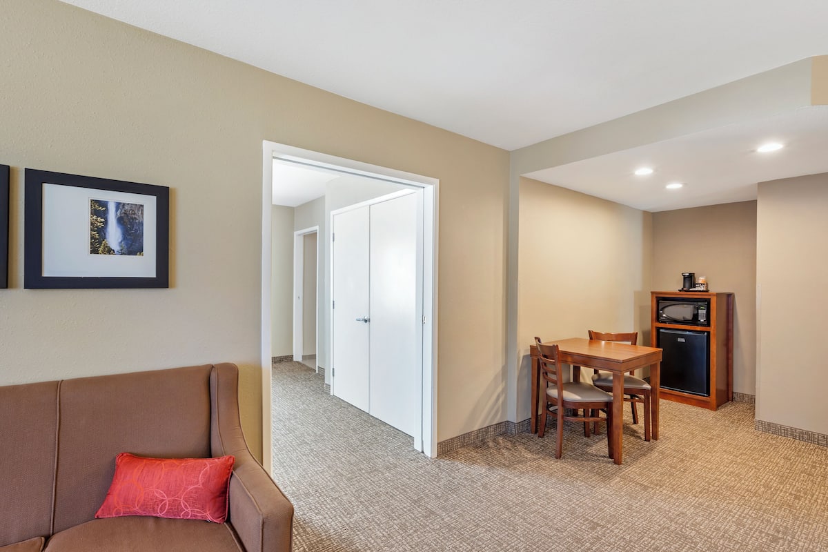 A hotel room with a brown sofa, a dining table with chairs, a microwave, and a refrigerator. There is a hallway leading to another room.