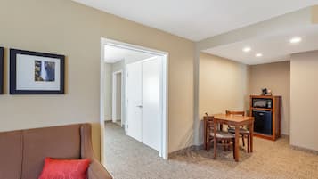 A hotel room with a brown sofa, a dining table with chairs, a microwave, and a refrigerator. There is a hallway leading to another room.