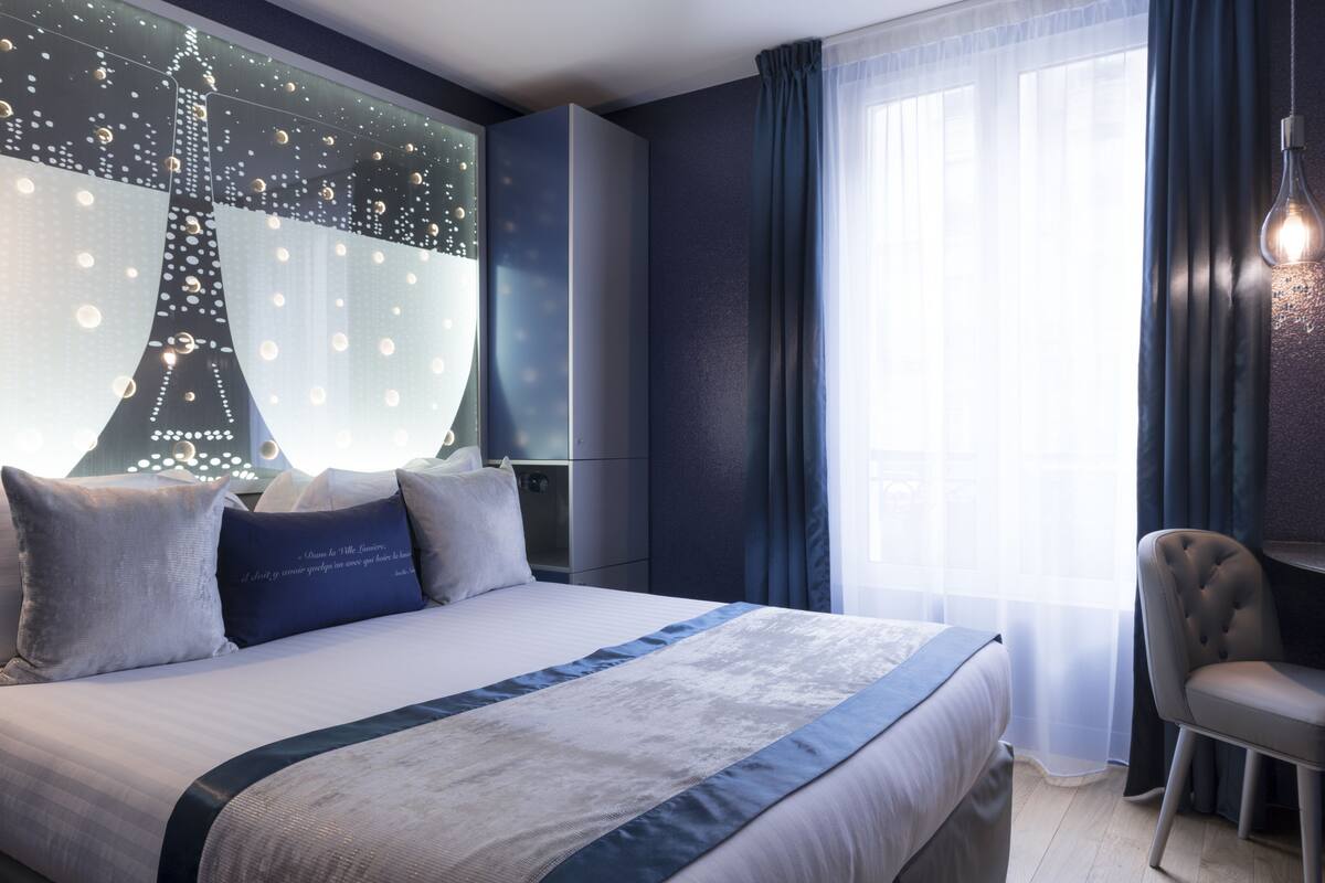 Une chambre d’hôtel moderne avec un grand lit, une vue sur la Tour Eiffel affichée sur le mur, une chaise et un bureau.