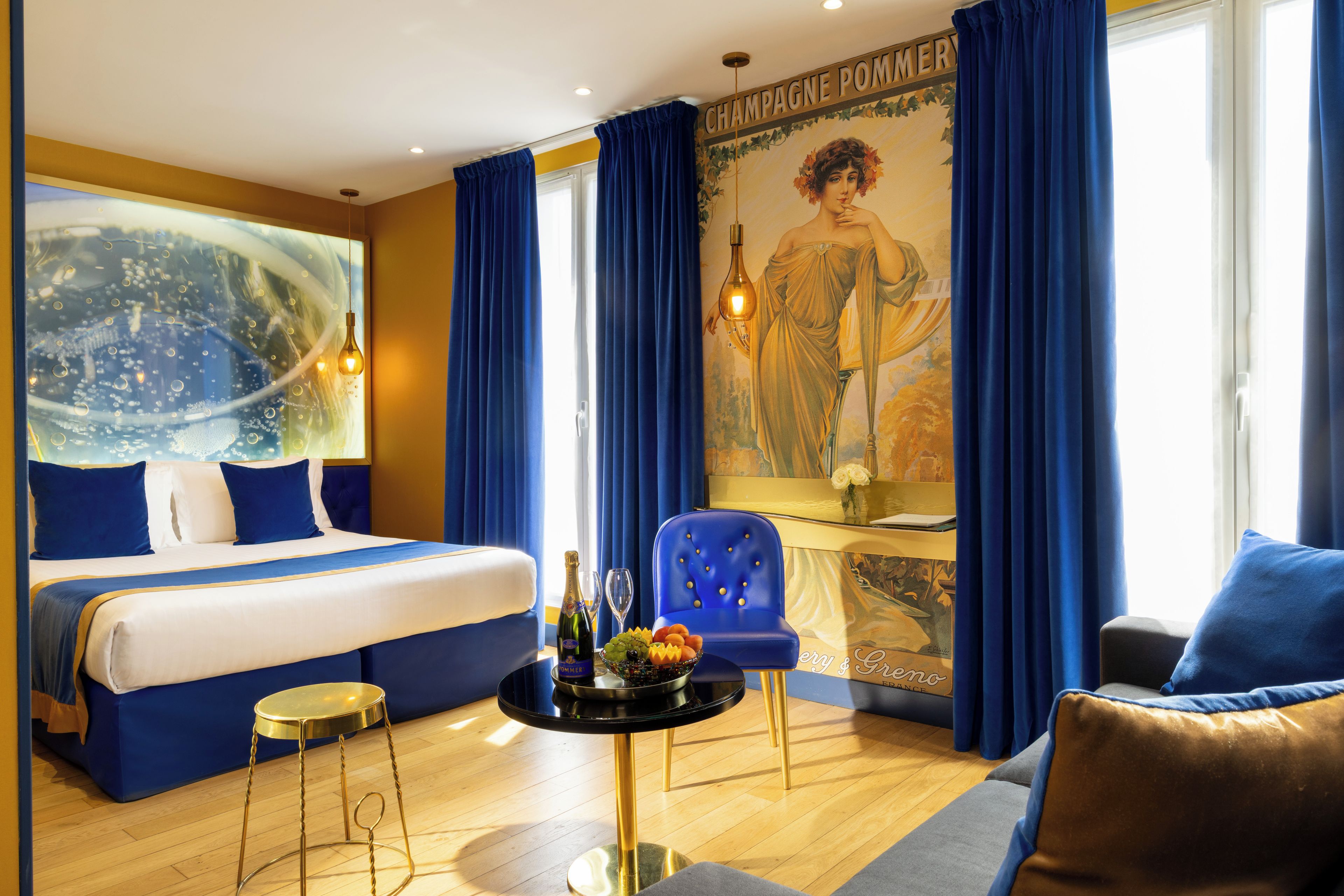 Une chambre d’hôtel avec un lit, une table ronde sur laquelle se trouve une bouteille de champagne, un fauteuil bleu et un grand tableau accroché au mur.