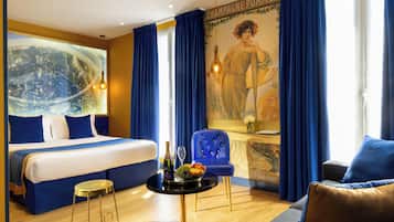 Une chambre d’hôtel avec un lit, une table ronde sur laquelle se trouve une bouteille de champagne, un fauteuil bleu et un grand tableau accroché au mur.