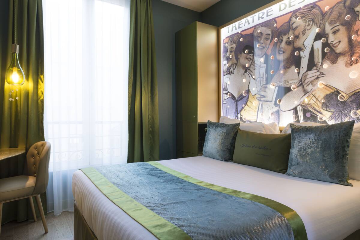 Une chambre d’hôtel avec un lit, un bureau et une chaise. Il y a une affiche au mur et une fenêtre avec des rideaux.