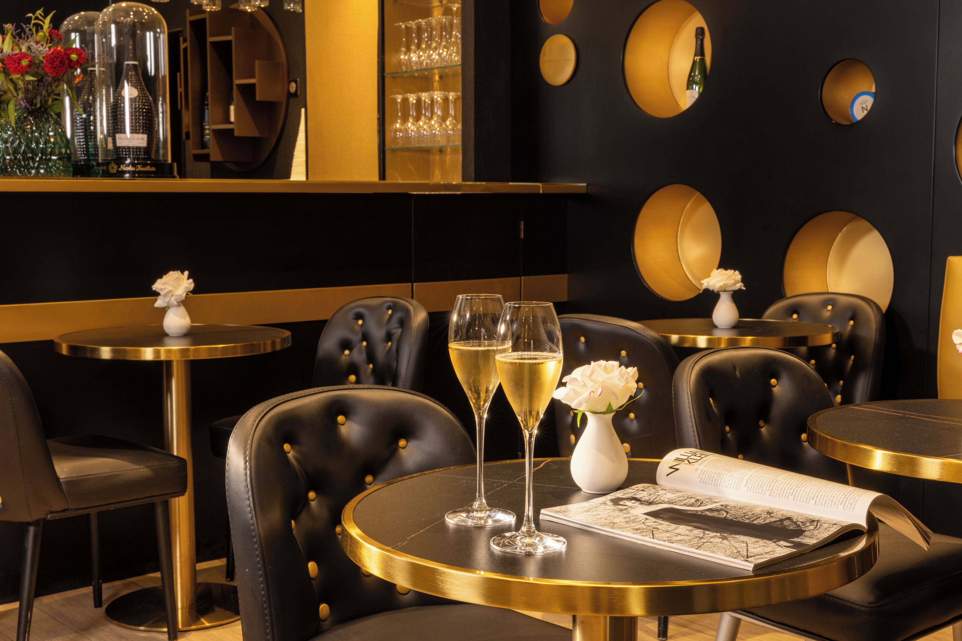 L’intérieur d’un restaurant présente des tables rondes, des chaises en cuir noir et un bar avec des verres et des bouteilles de vin.