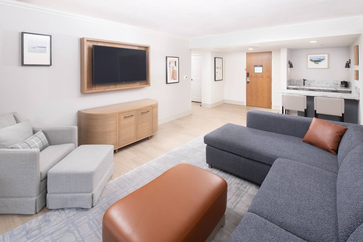A modern living room with a grey sofa, a brown ottoman, and a grey couch. There is a wooden cabinet, a TV mounted on the wall, and a kitchen area in the background.