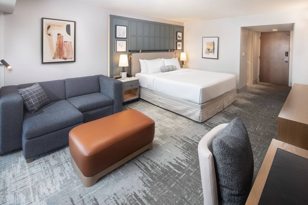 A hotel room with a bed, a grey sofa, a brown ottoman, a desk, and a chair.
