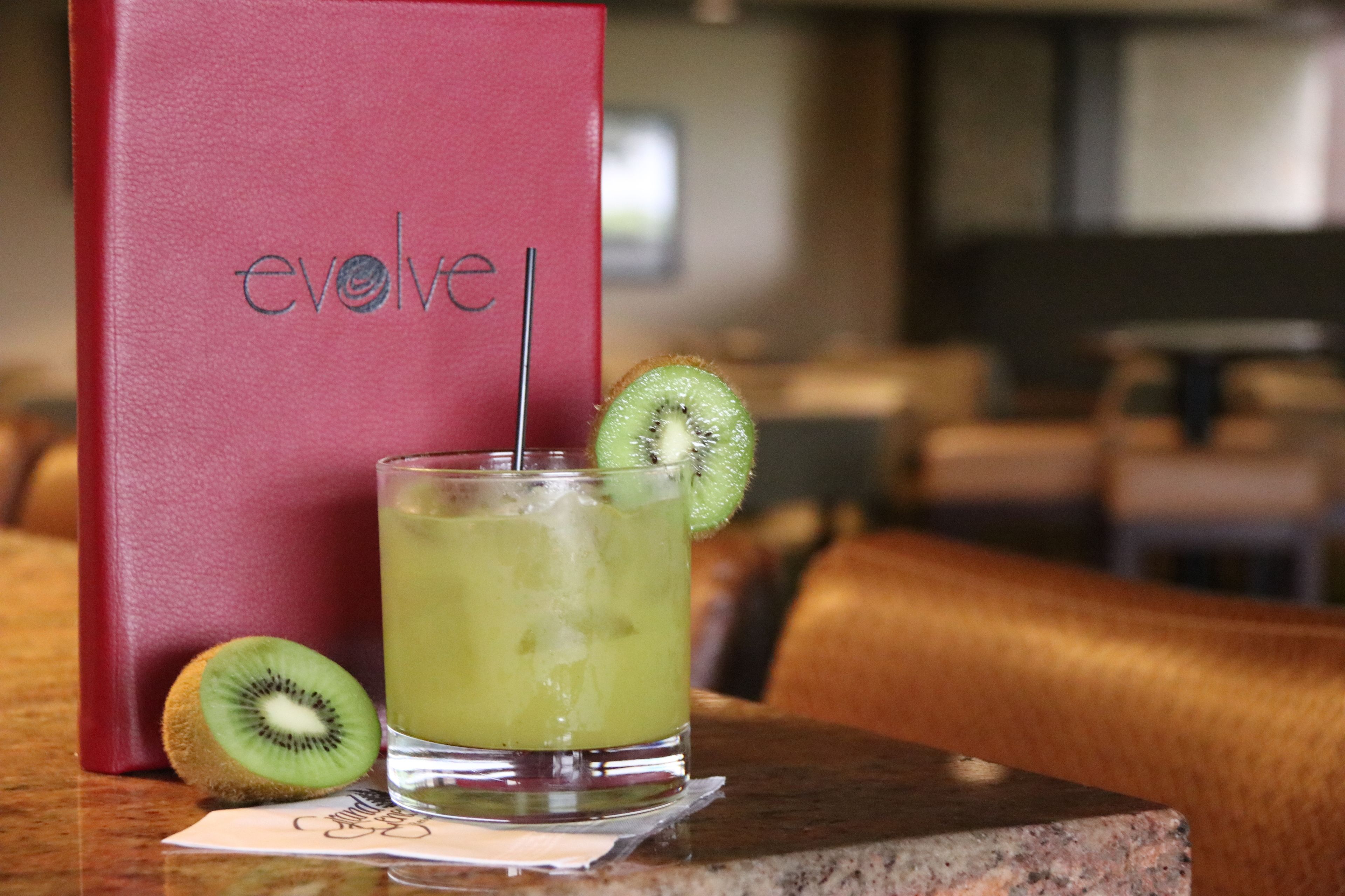 A glass of green beverage with a kiwi slice, a red book titled 'evolve', and a napkin with a drawing on a table.