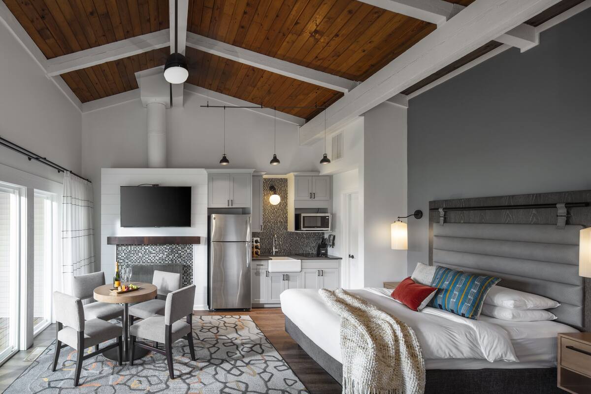 A modern hotel room with a large bed, a dining area, and a kitchenette.