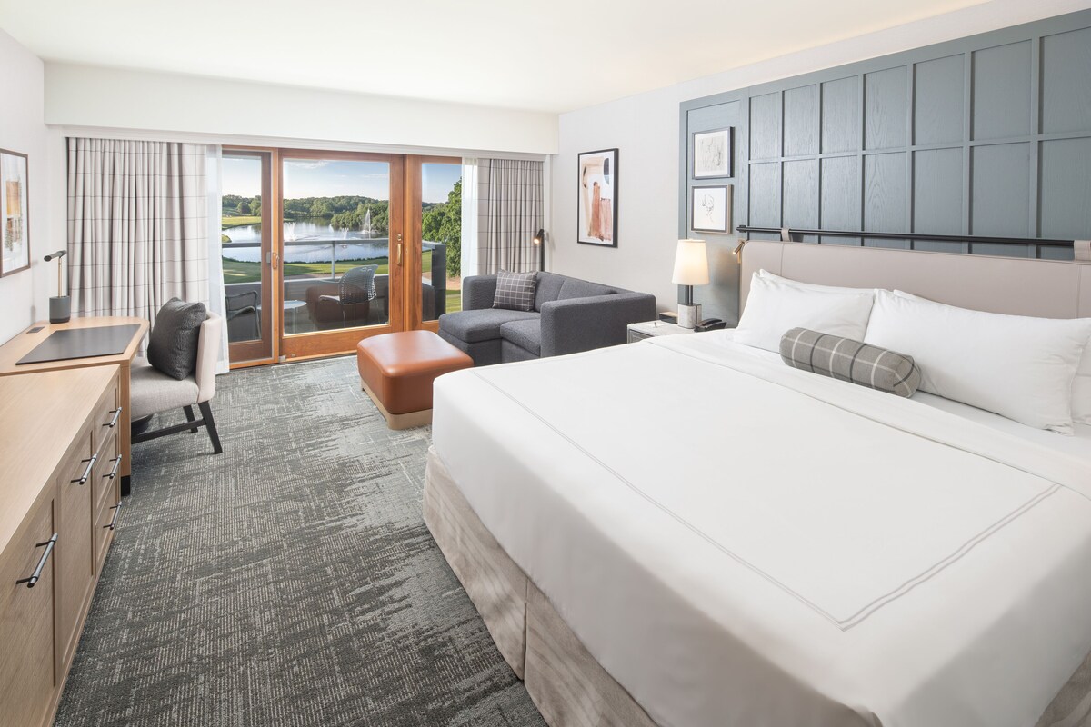A hotel room with a large bed, a desk, a chair, and a view of a lake through the glass doors.