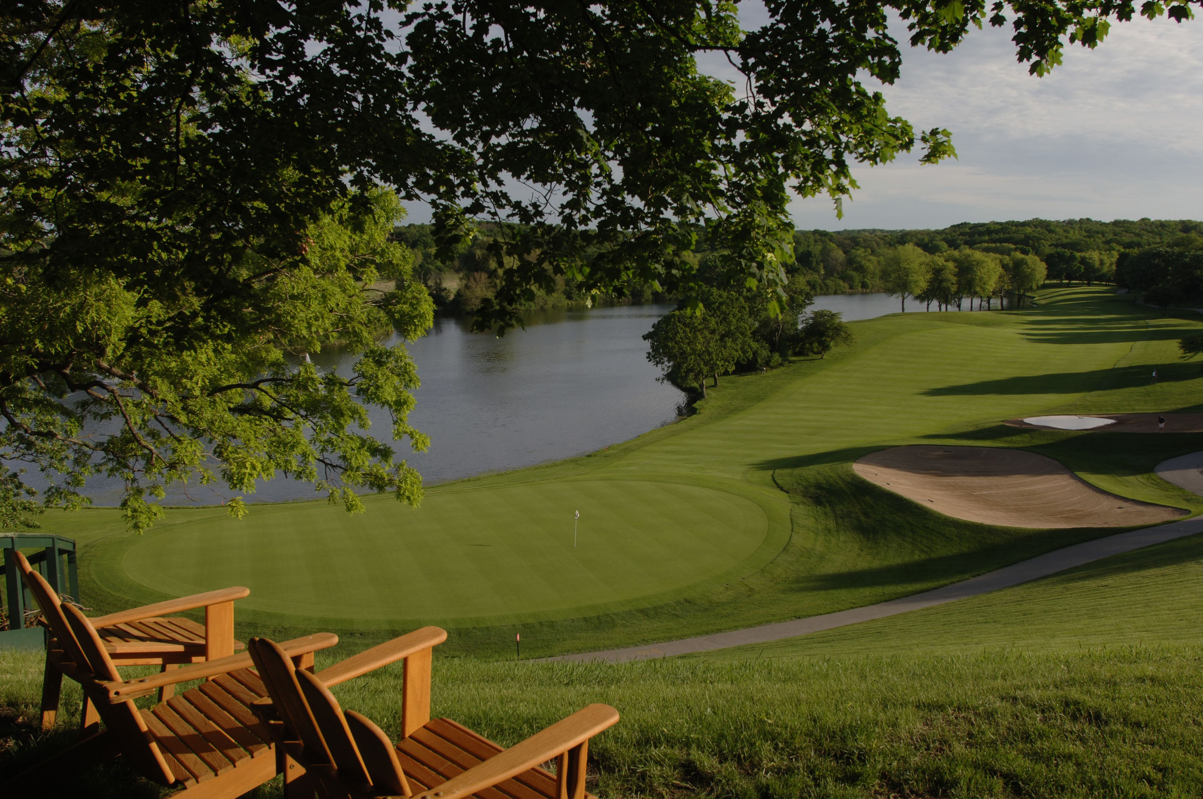 A golf course with a water body, trees, and wooden chairs.