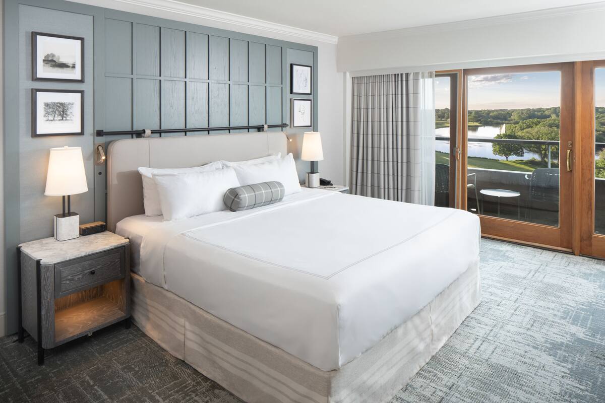 A large bed with white linens, a nightstand with a lamp, and a view of a lake through a sliding glass door.