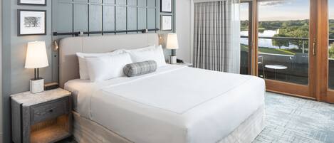 Una cama grande con sábanas blancas, una mesita de noche con lámpara, y vista a un lago a través de una puerta corrediza de vidrio.