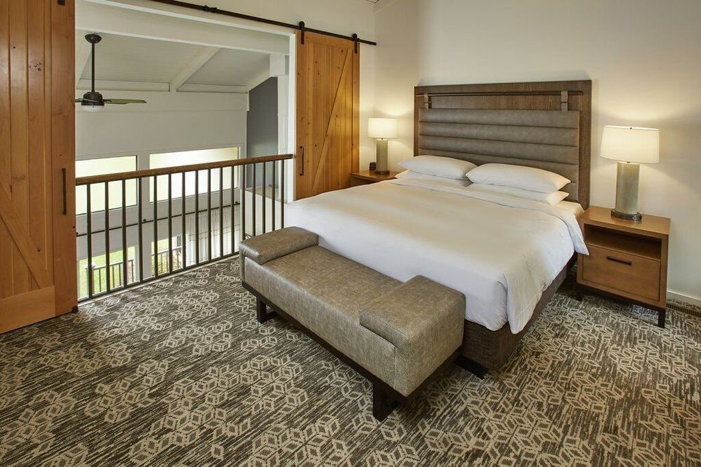 A bedroom with a large bed, two bedside tables with lamps, a bench, a patterned carpet, and a balcony with a railing.