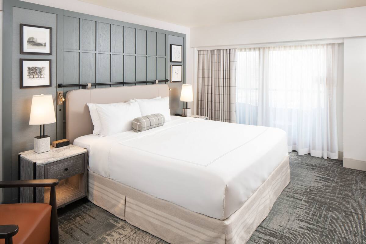 A large bed with white linens, two bedside tables with lamps, a wooden nightstand, a chair, and a view of a window with curtains.
