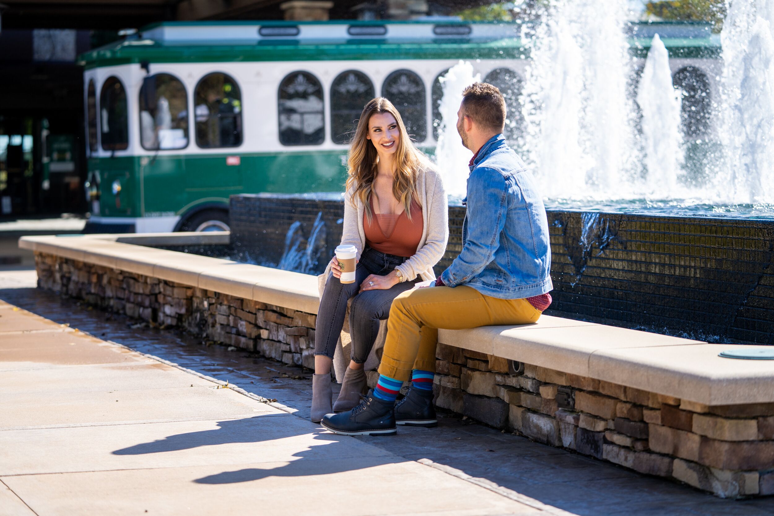 A couple sitting on a stone bench near a fountain, with a green bus in the background.
