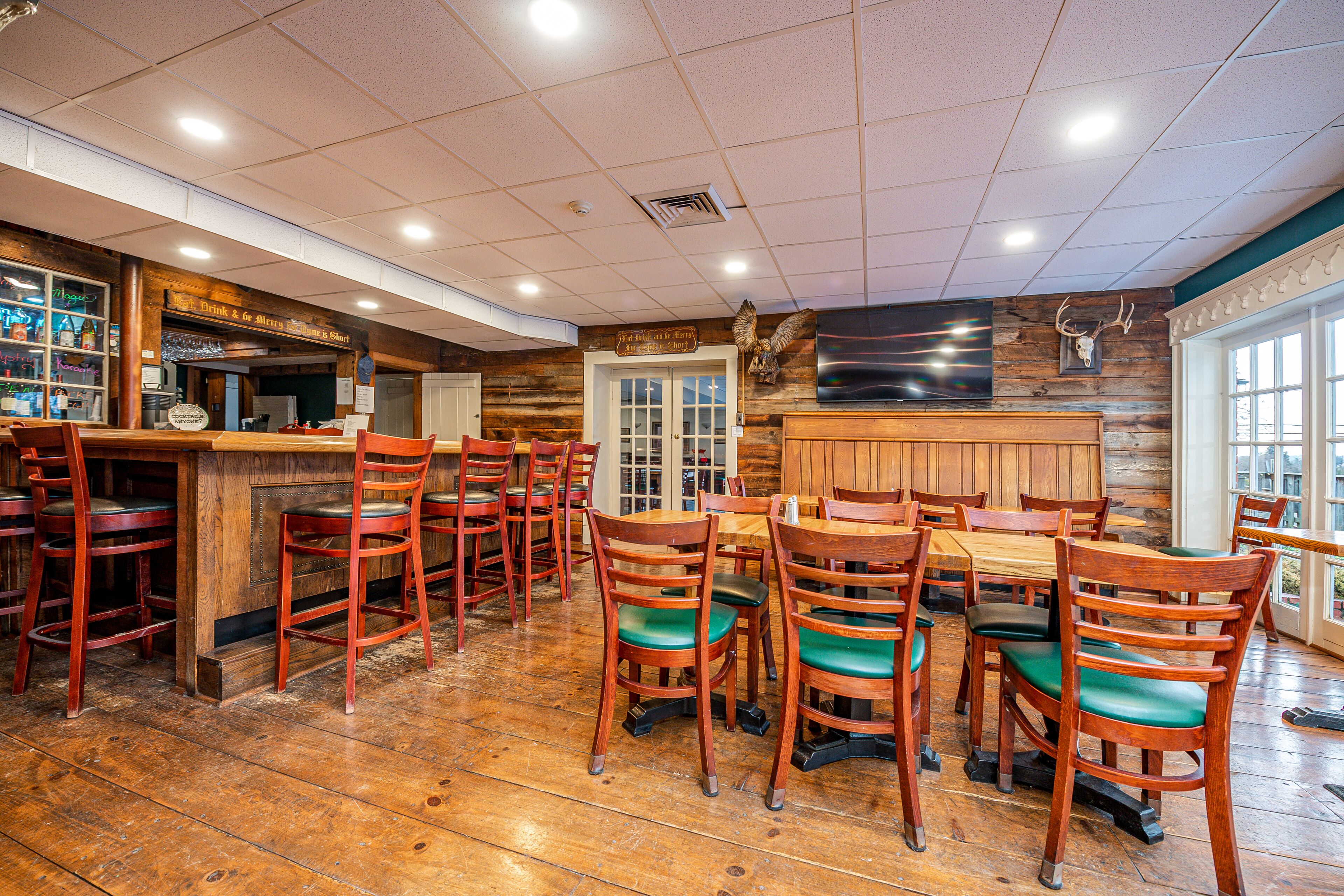 Un restaurante con suelo de madera, mesas y sillas de madera, una zona de bar y un televisor montado.