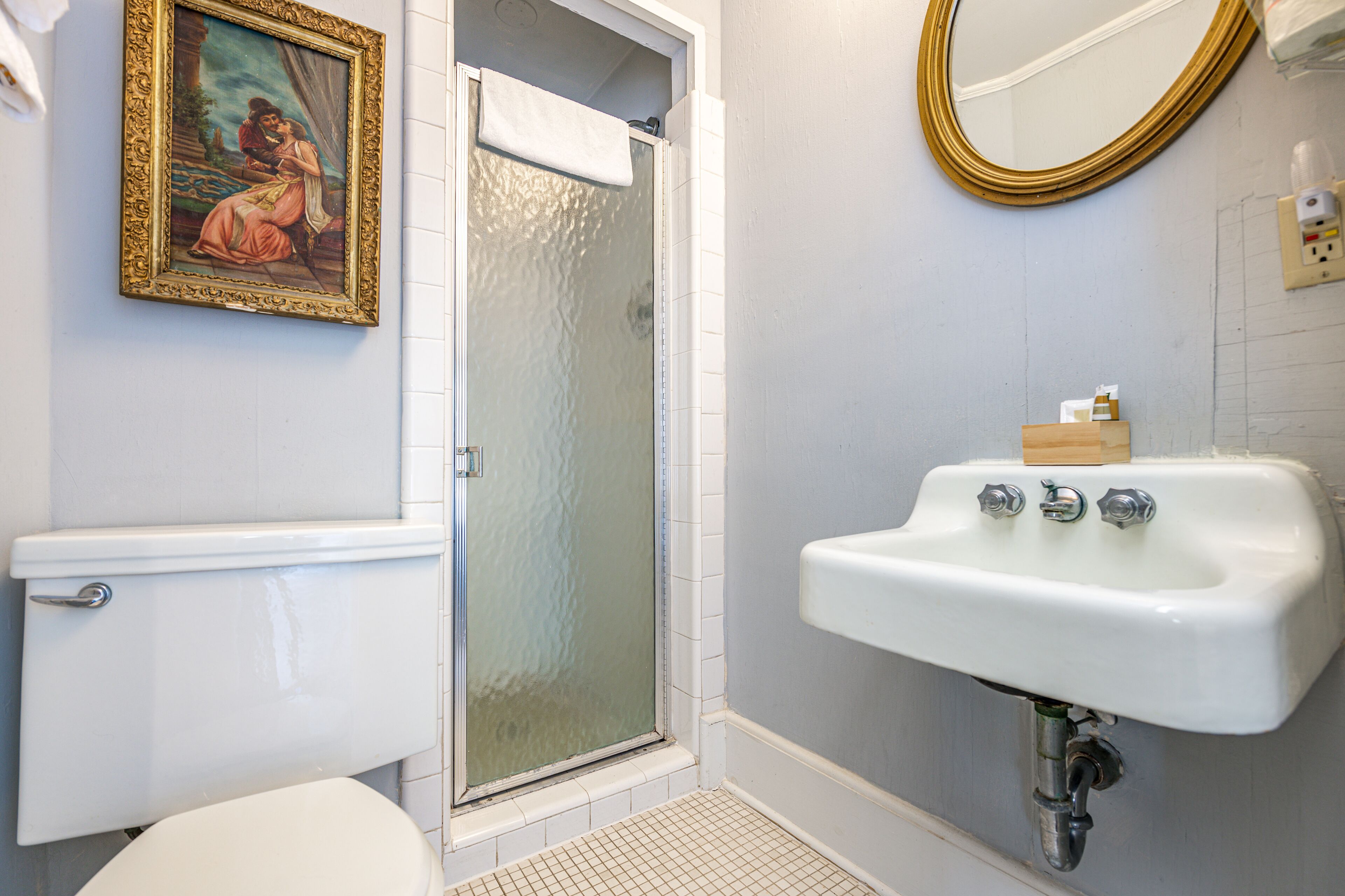 Un baño con inodoro, lavamanos, ducha con puerta de vidrio y un cuadro en la pared.