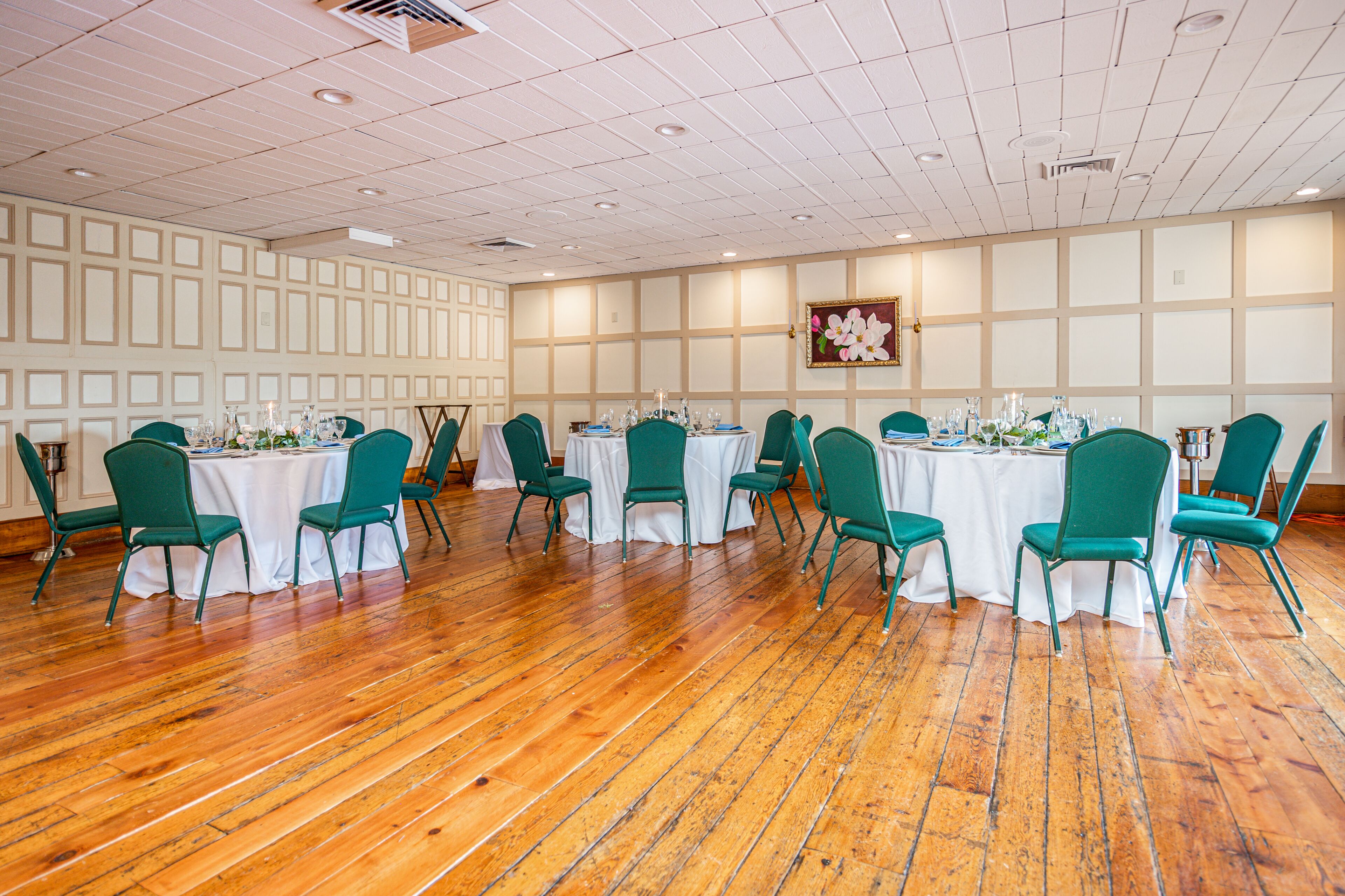 A conference room with wooden flooring, green chairs, white tables, and a floral painting on the wall.