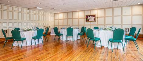 A conference room with wooden flooring, green chairs, white tables, and a floral painting on the wall.