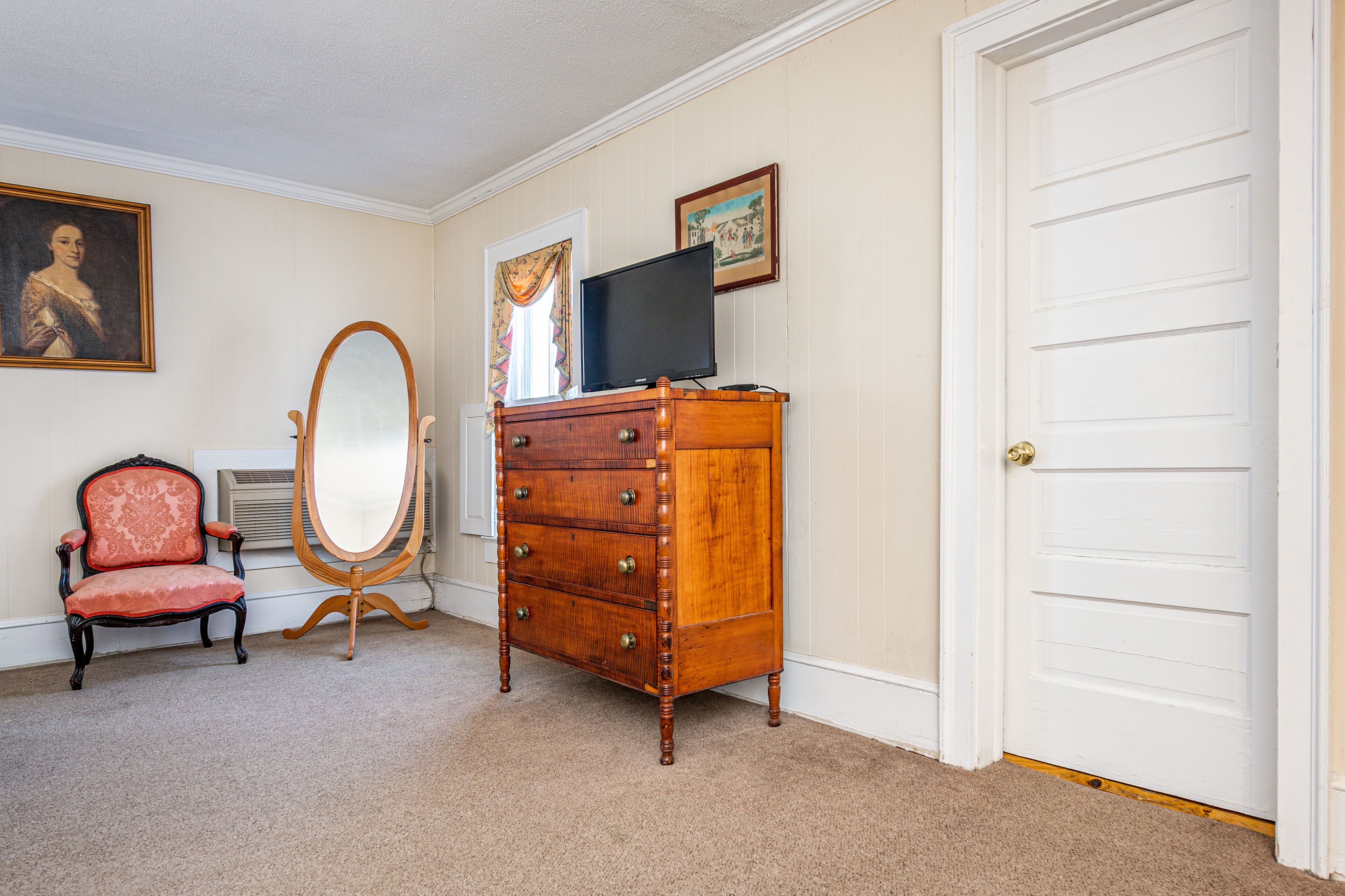 A room with a wooden dresser, a chair, a mirror, a television, and a door.