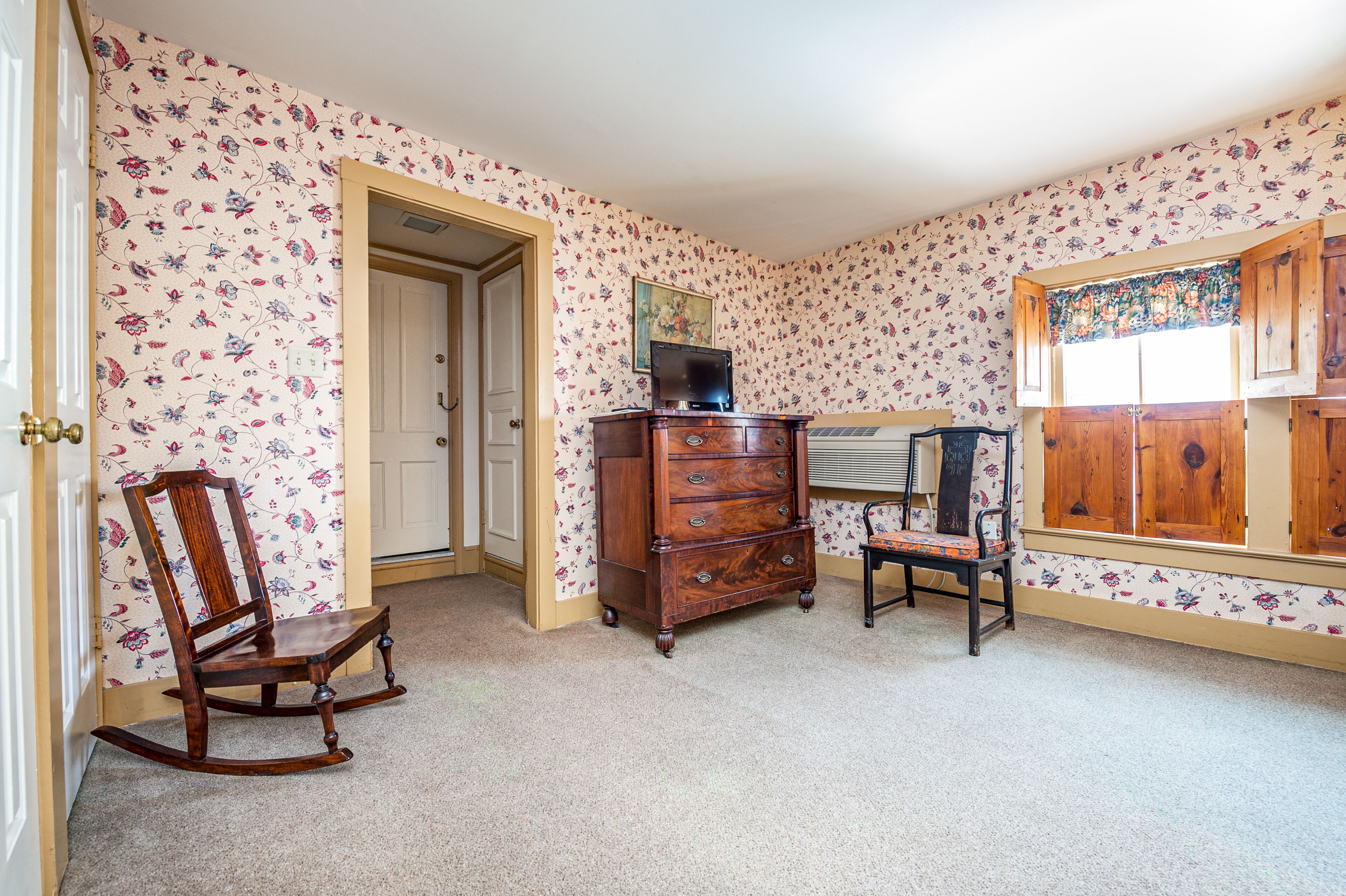 A room with floral wallpaper, a wooden rocking chair, a dresser, and a window with shutters.