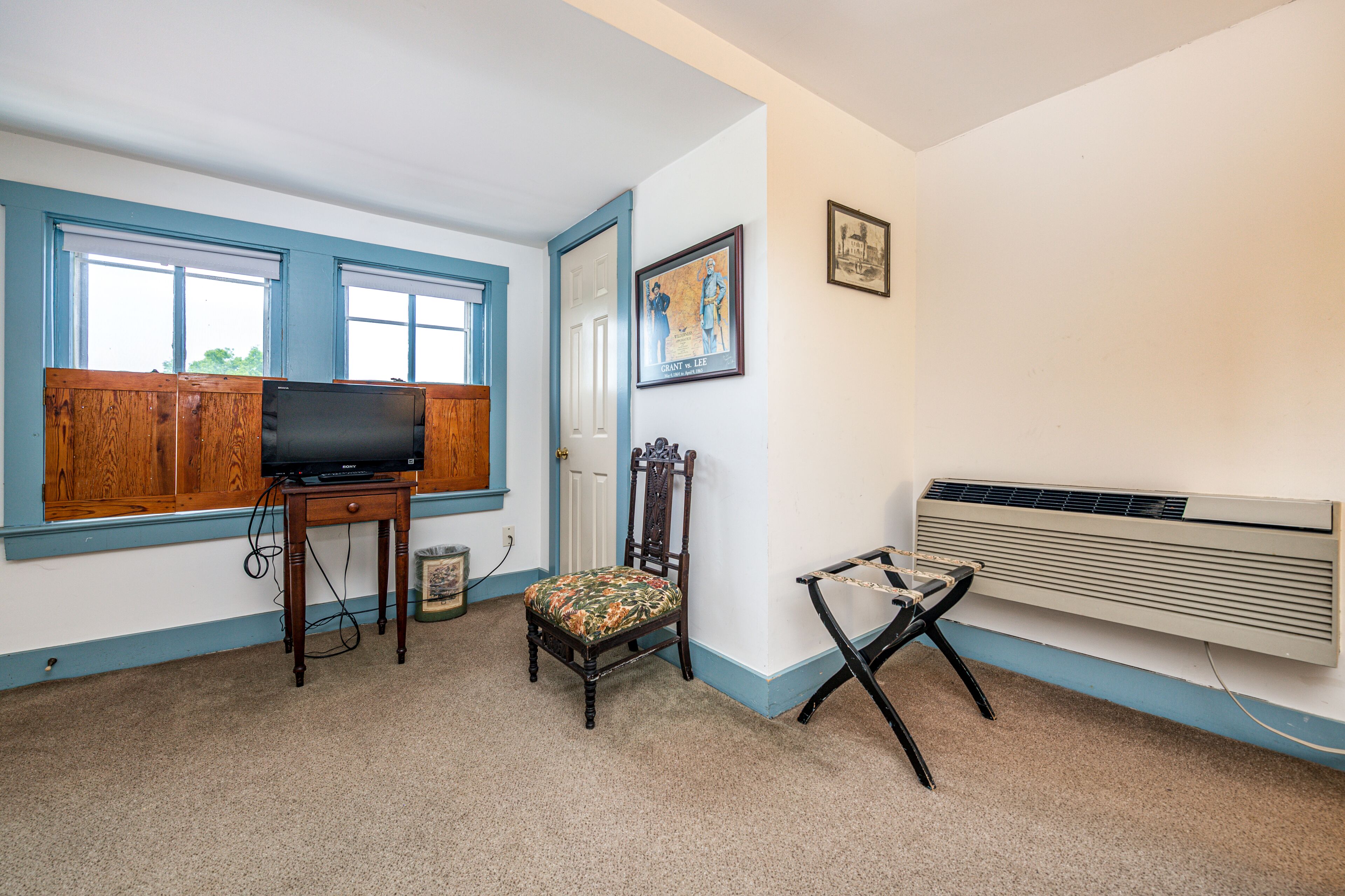 Una habitación con una televisión sobre una mesa de madera, una silla con cojín floral, un radiador y una puerta que da a otra habitación.