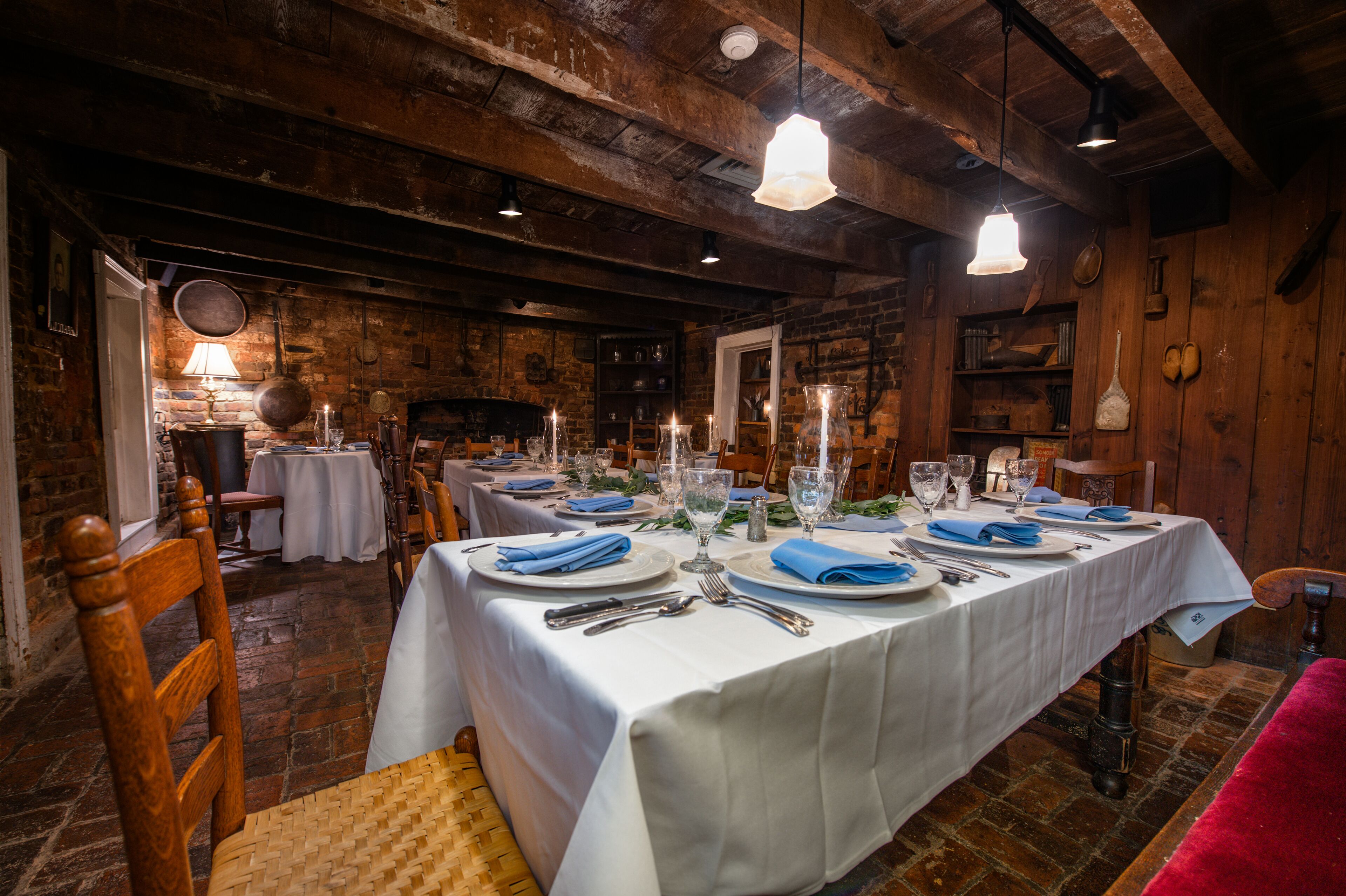 Une salle à manger rustique aux murs en bois et aux poutres apparentes, avec plusieurs tables dressées pour le repas, recouvertes de nappes blanches et agrémentées de serviettes bleues.