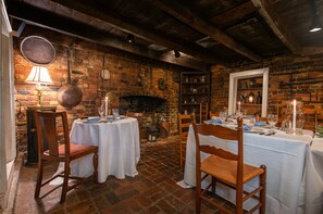 A rustic dining area with brick walls, wooden beams, and a fireplace. Tables are set with white tablecloths and candles.