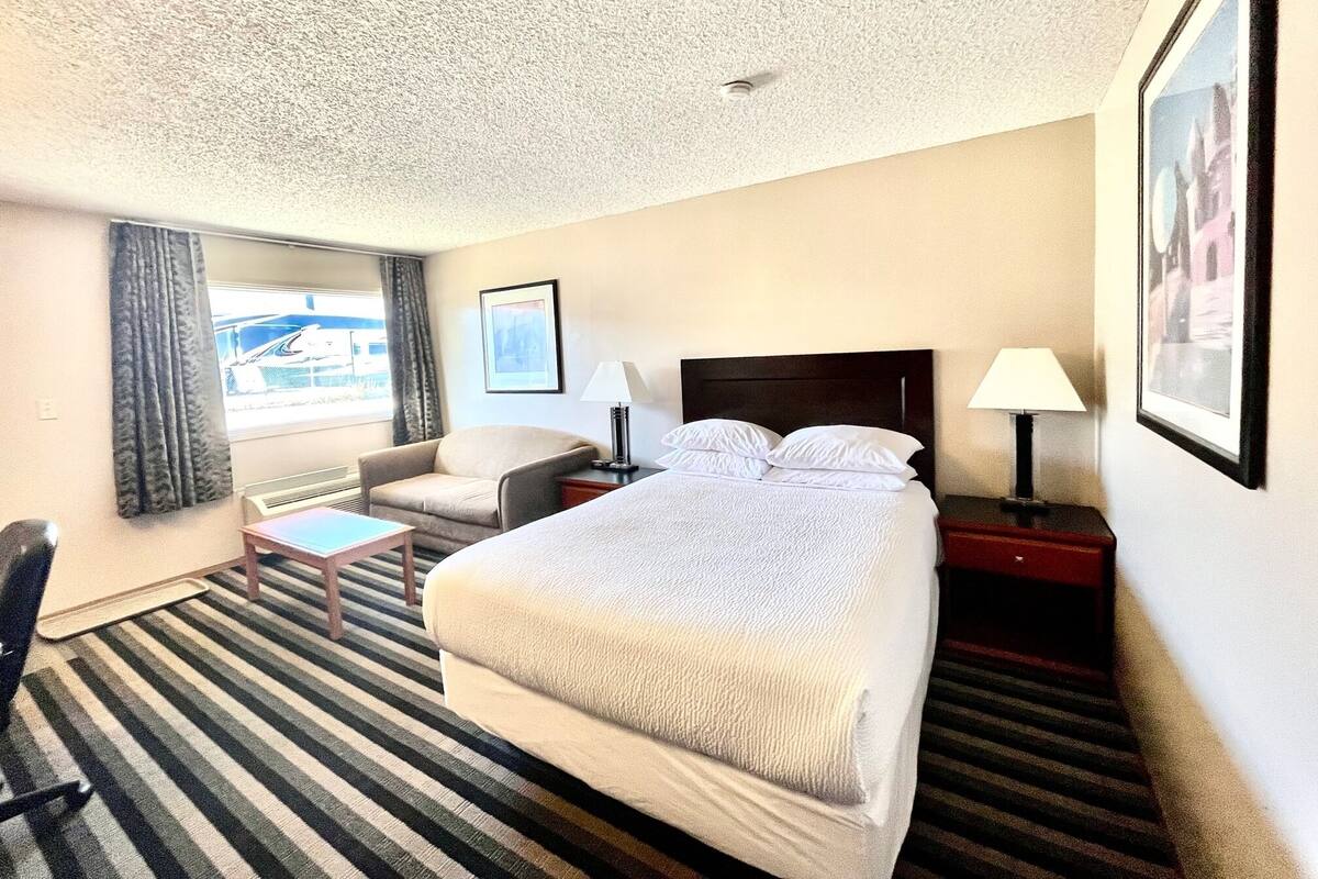 A hotel room with a bed, a chair, a desk, a lamp, and a window with curtains.