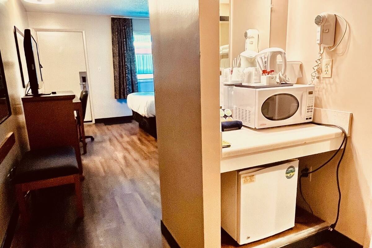 A hotel room with a wooden floor, a desk with a chair, a bed, a microwave, a hairdryer, and a mirror with lights.