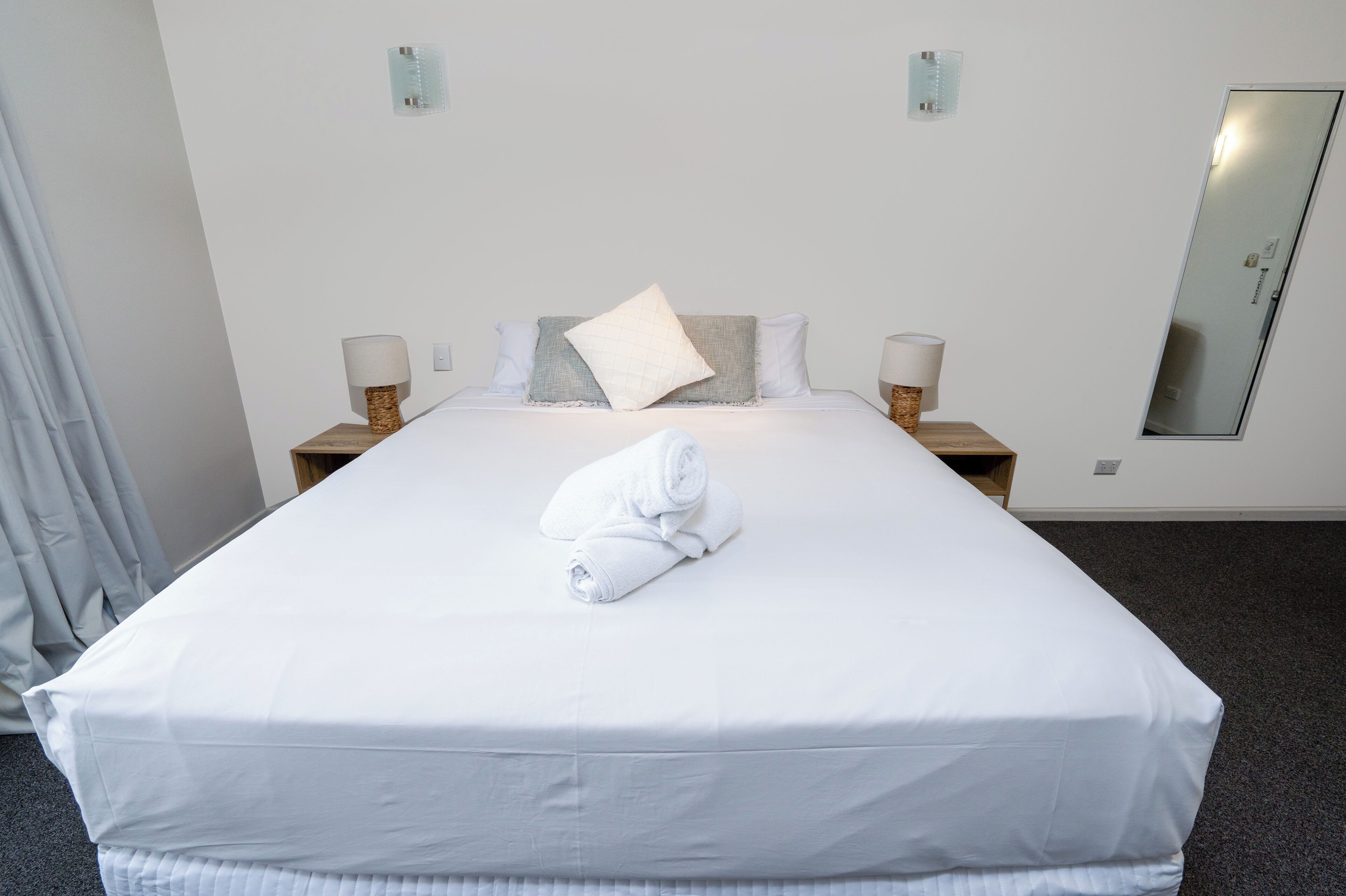 A neatly made bed with a rolled towel, two bedside lamps, and a mirror on the wall.