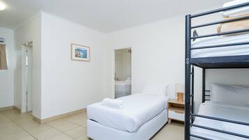 A single bed with a white mattress and matching pillows, a wooden nightstand with a lamp, and a bunk bed with a towel on the top bunk.