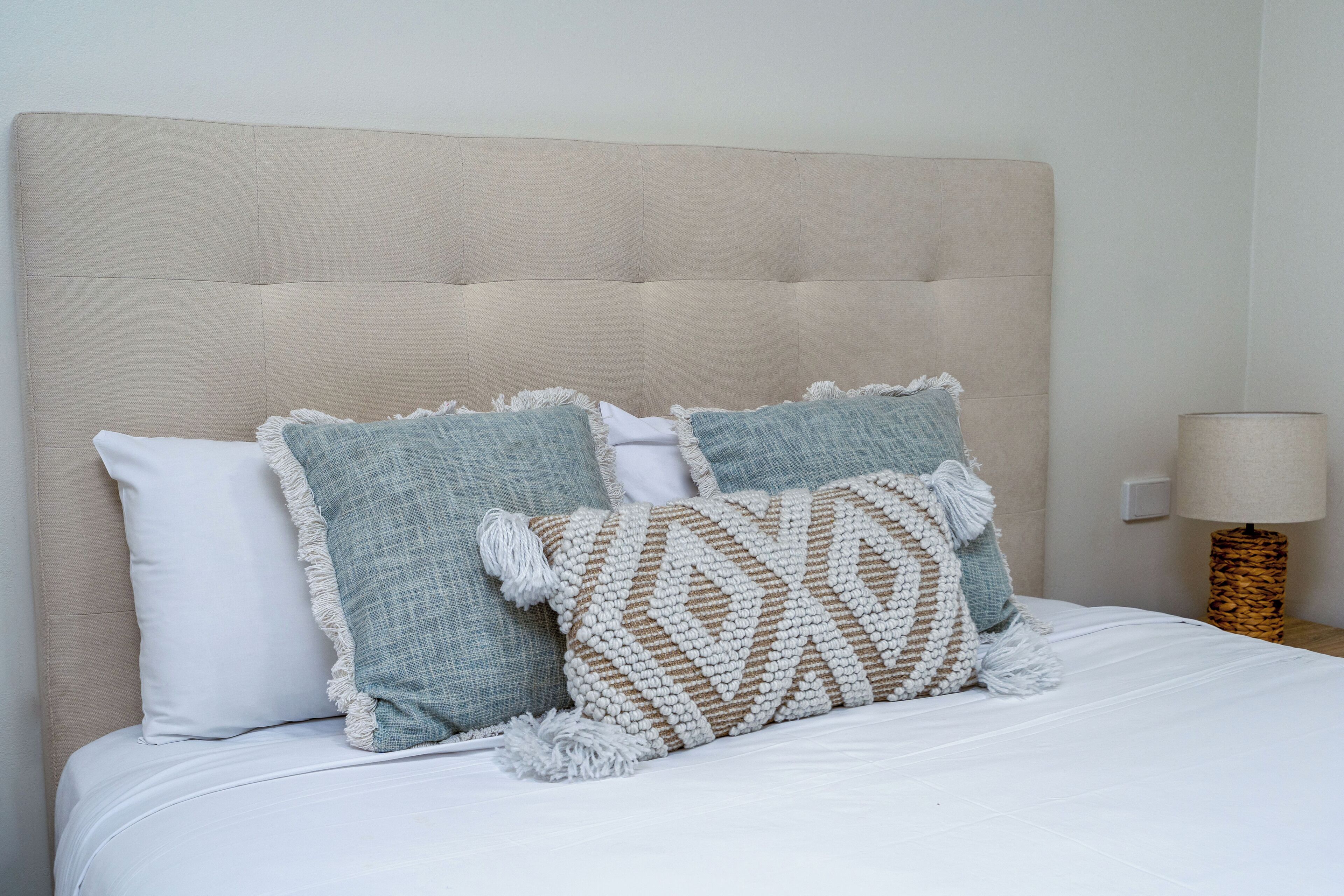 A bed with a beige upholstered headboard, two pillows with fringe details, and a woven throw blanket.