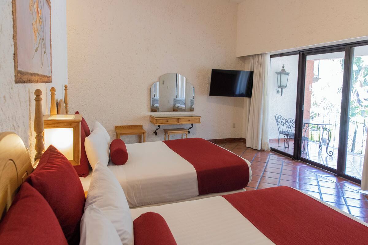Habitación de hotel con dos camas, televisión, escritorio y balcón con vistas.