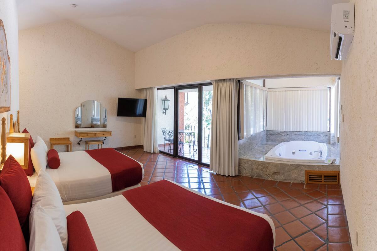 Habitación de hotel con dos camas, televisión, baño con bañera y balcón con cortinas.
