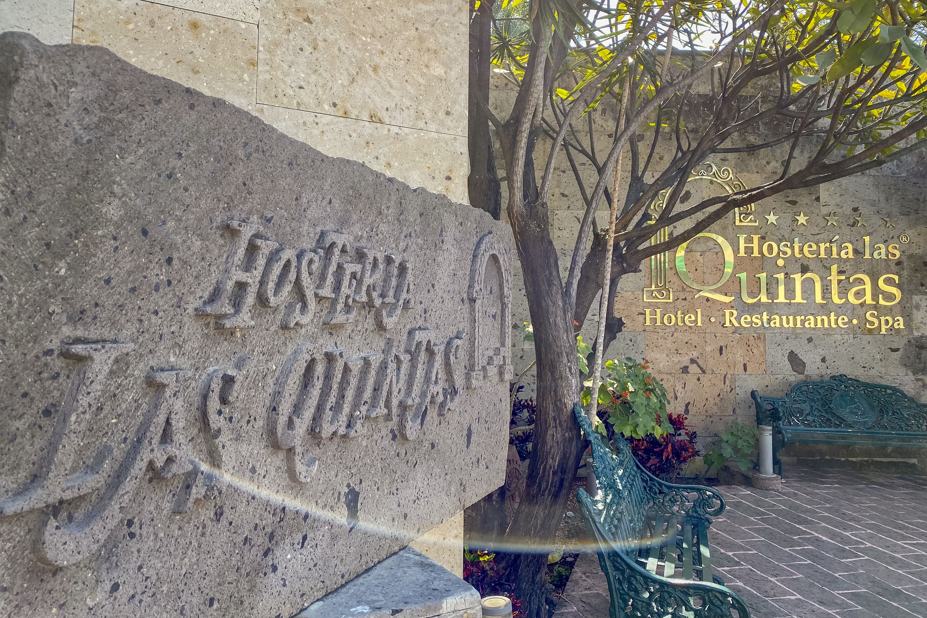 Un letrero de piedra con la inscripción "Hosteria las Quintas", un banco verde y un árbol con hojas.