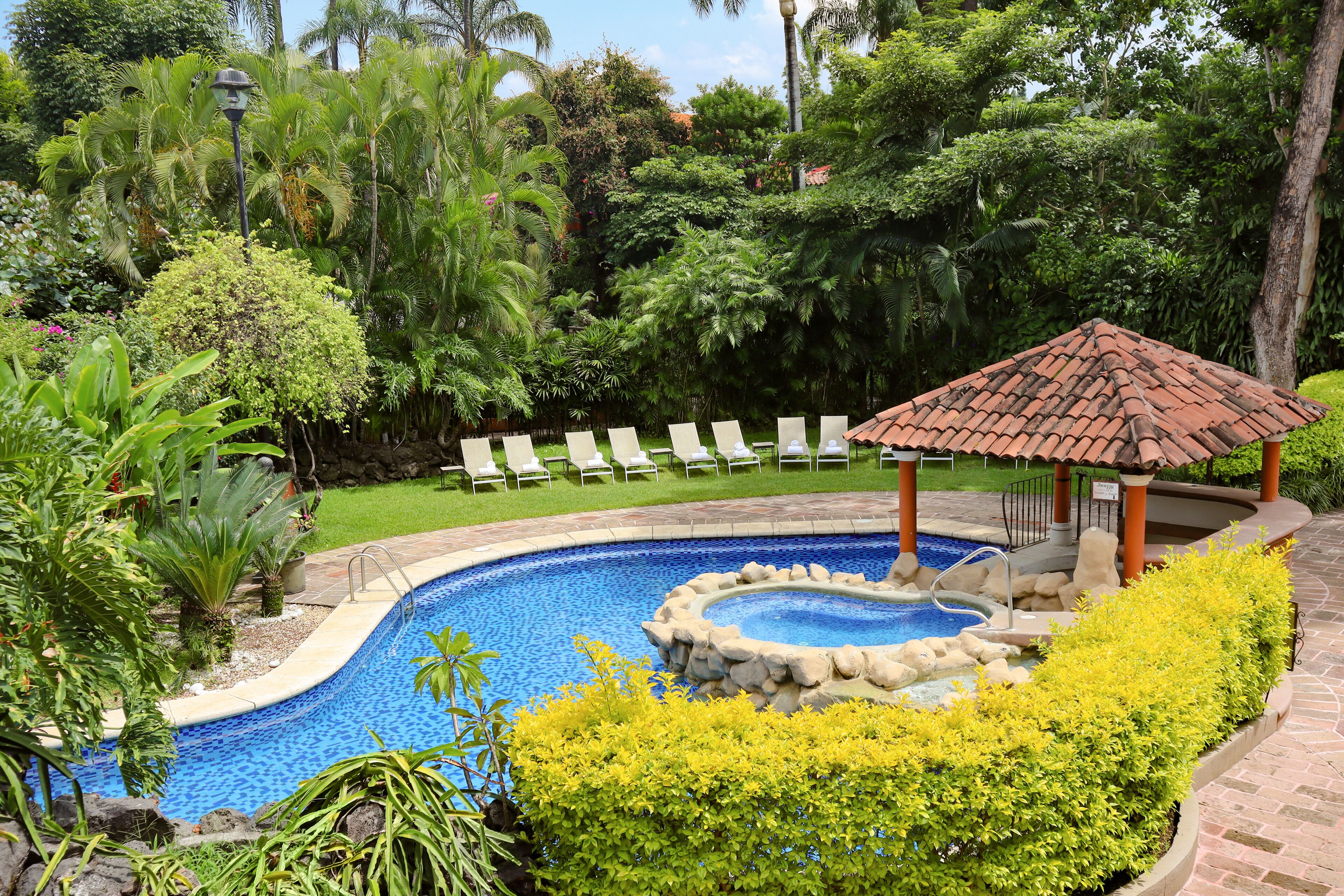 Uma piscina com uma banheira de hidromassagem circular, rodeada por vegetação exuberante e um gazebo com telhado em azulejos.
