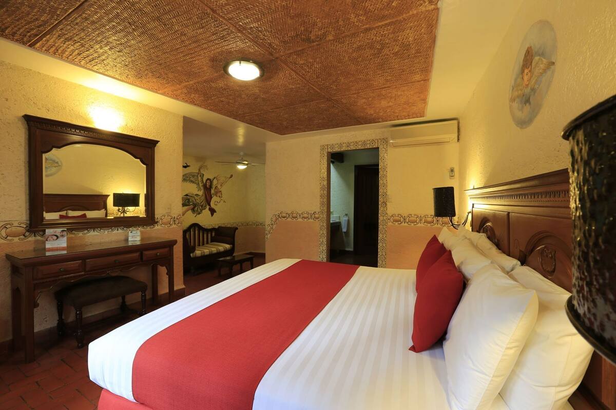 Habitación de hotel con una cama grande, un escritorio con silla, un espejo y un ventilador de techo.