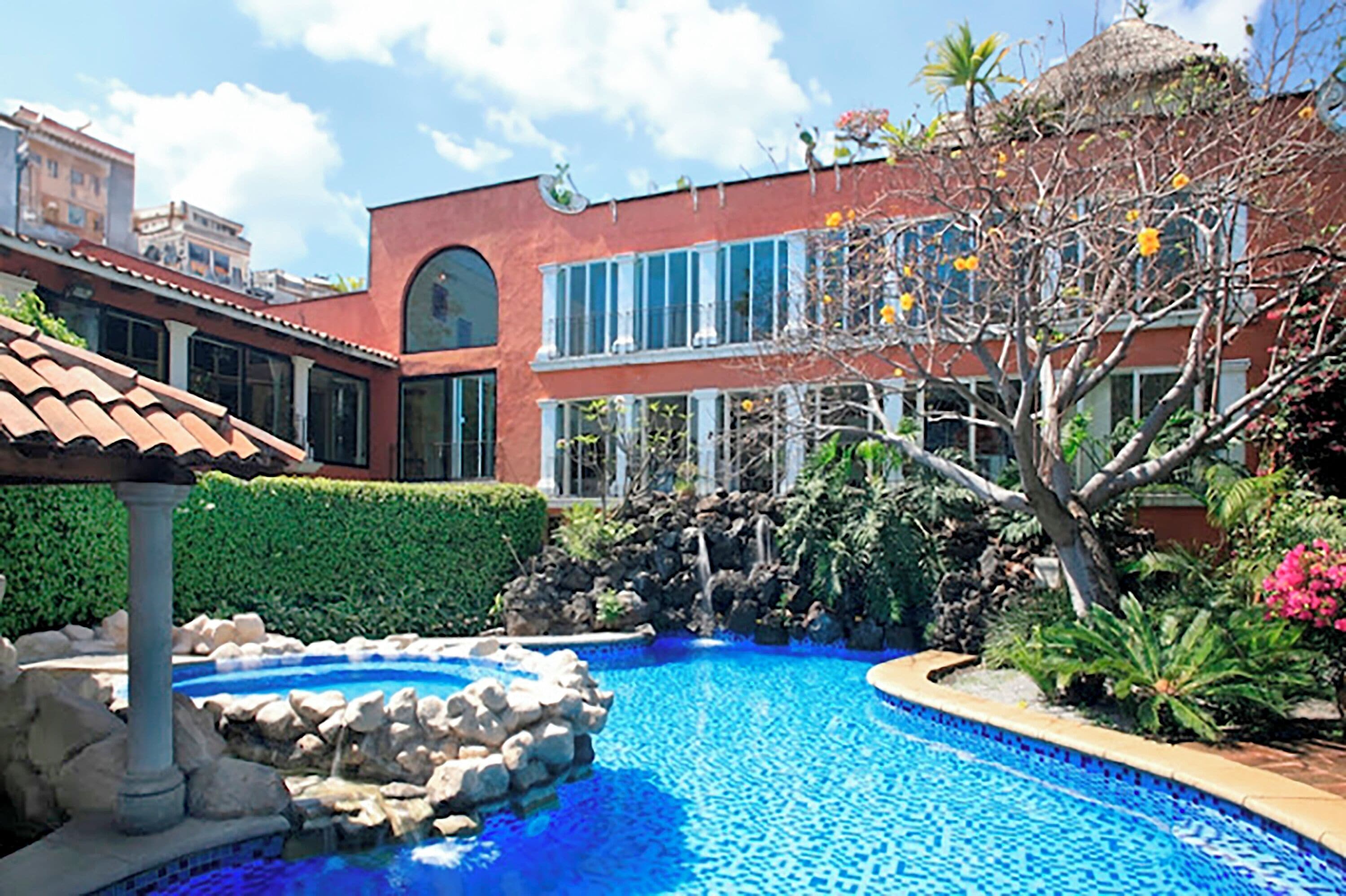 A swimming pool with a fountain, surrounded by greenery and a building with large windows.