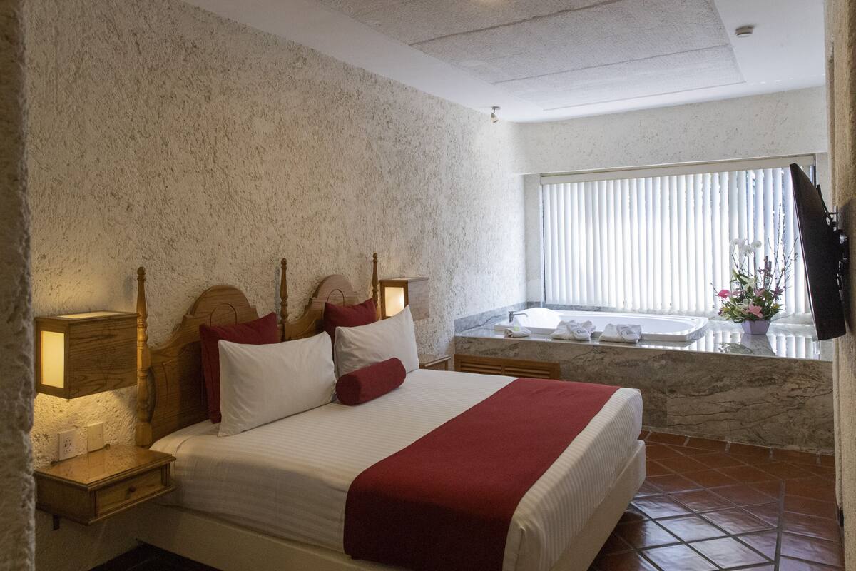 Habitación de hotel con una cama grande, cabecera de madera, lámparas de noche, tocador con encimera de mármol y una ventana con persianas.