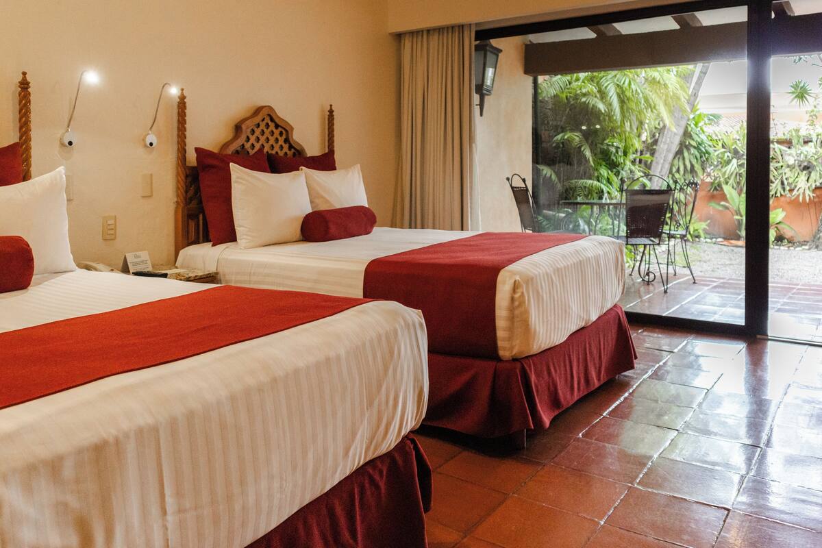 Habitación de hotel con dos camas, cada una con ropa de cama roja, cabeceros de madera y mesitas de noche con lámparas.