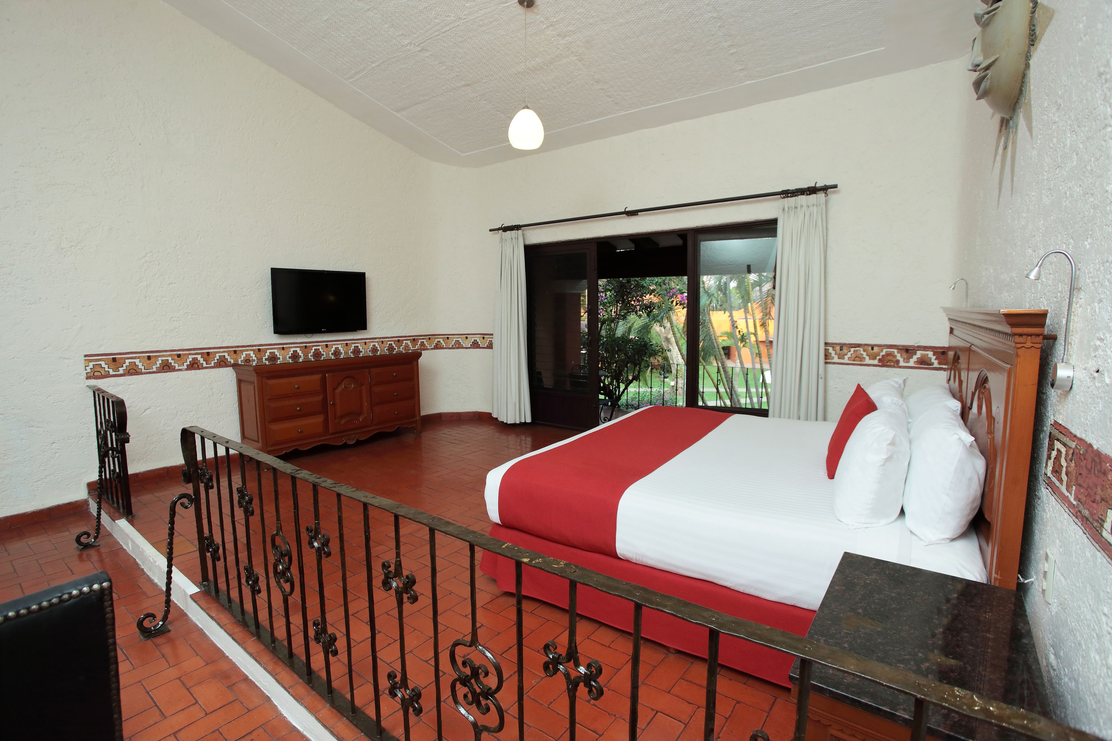 Habitación de hotel con cama, televisión, cómoda de madera, balcón con vistas y suelo de baldosas.