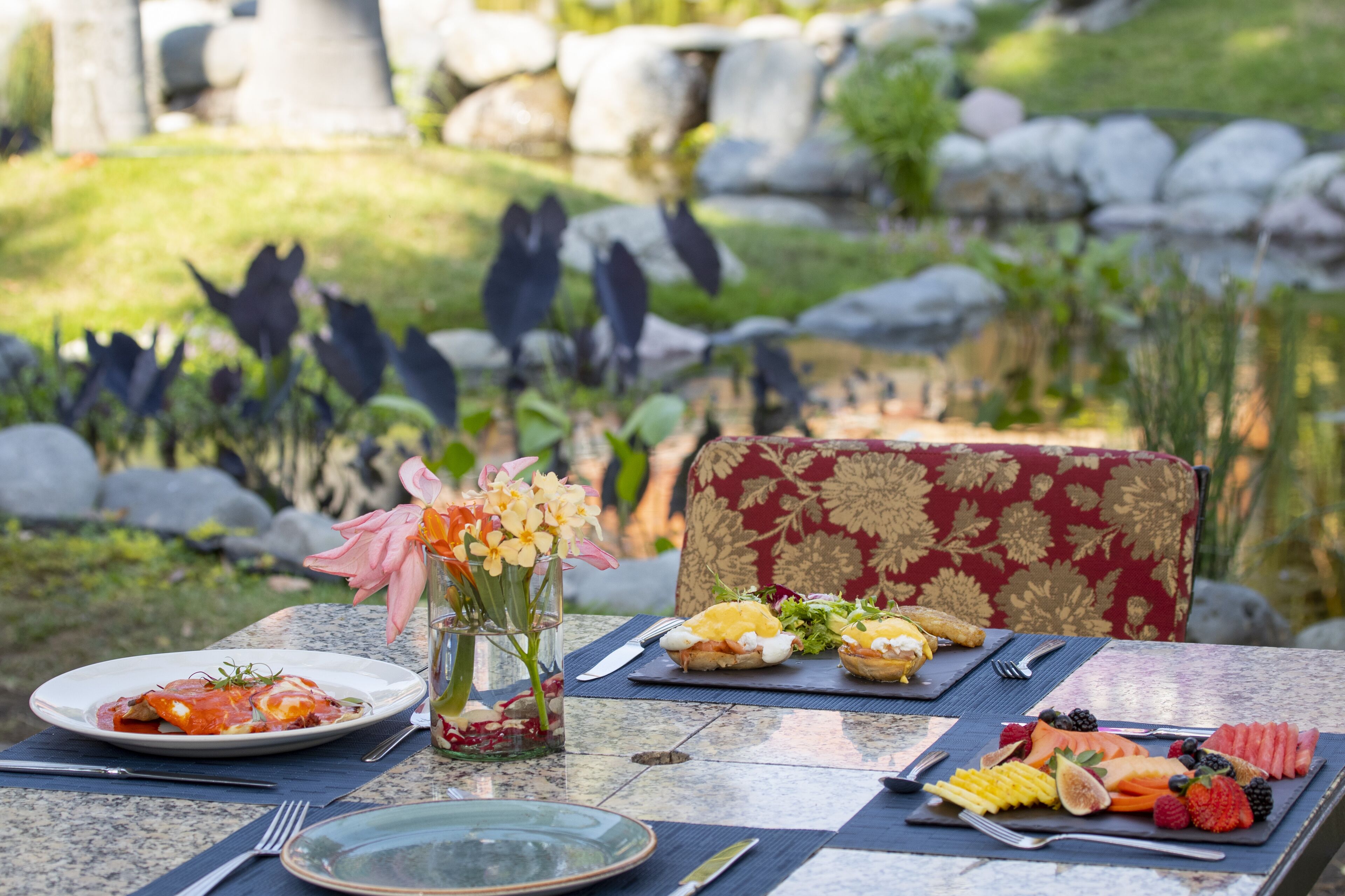 Mesa de comedor al aire libre con platos de comida, un florero y una silla con estampado floral.
