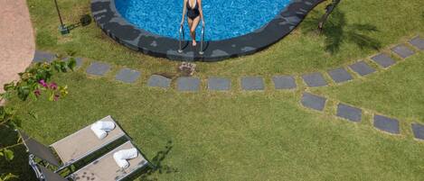 Una piscina con borde curvo, rodeada por un borde de piedra y césped. Hay sillones de descanso con toallas.