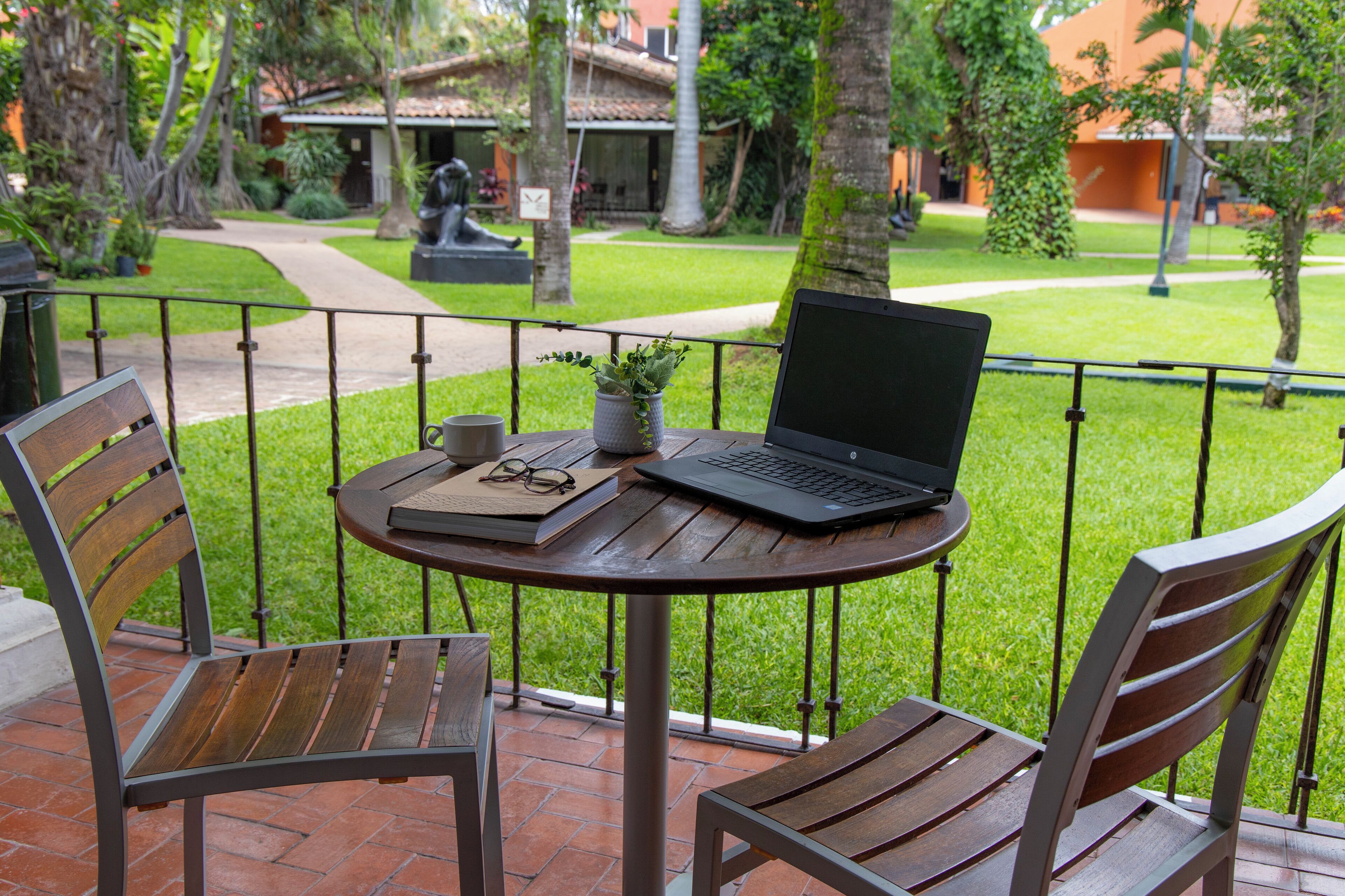 Una mesa redonda de madera con una computadora portátil, un libro y una taza en un balcón con barandillas de metal.