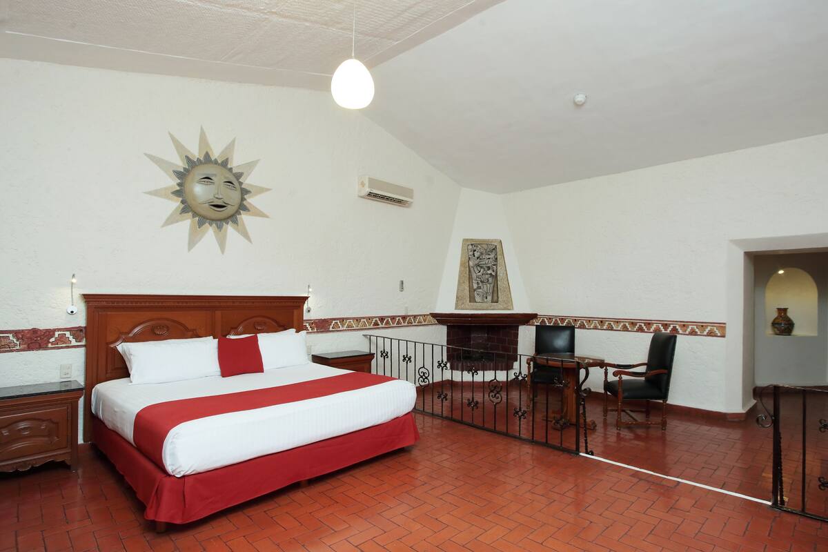 Habitación de hotel con una cama grande, cabecera de madera, colcha roja y blanca, decoración en forma de sol en la pared, chimenea, escritorio con silla y suelo de baldosas.