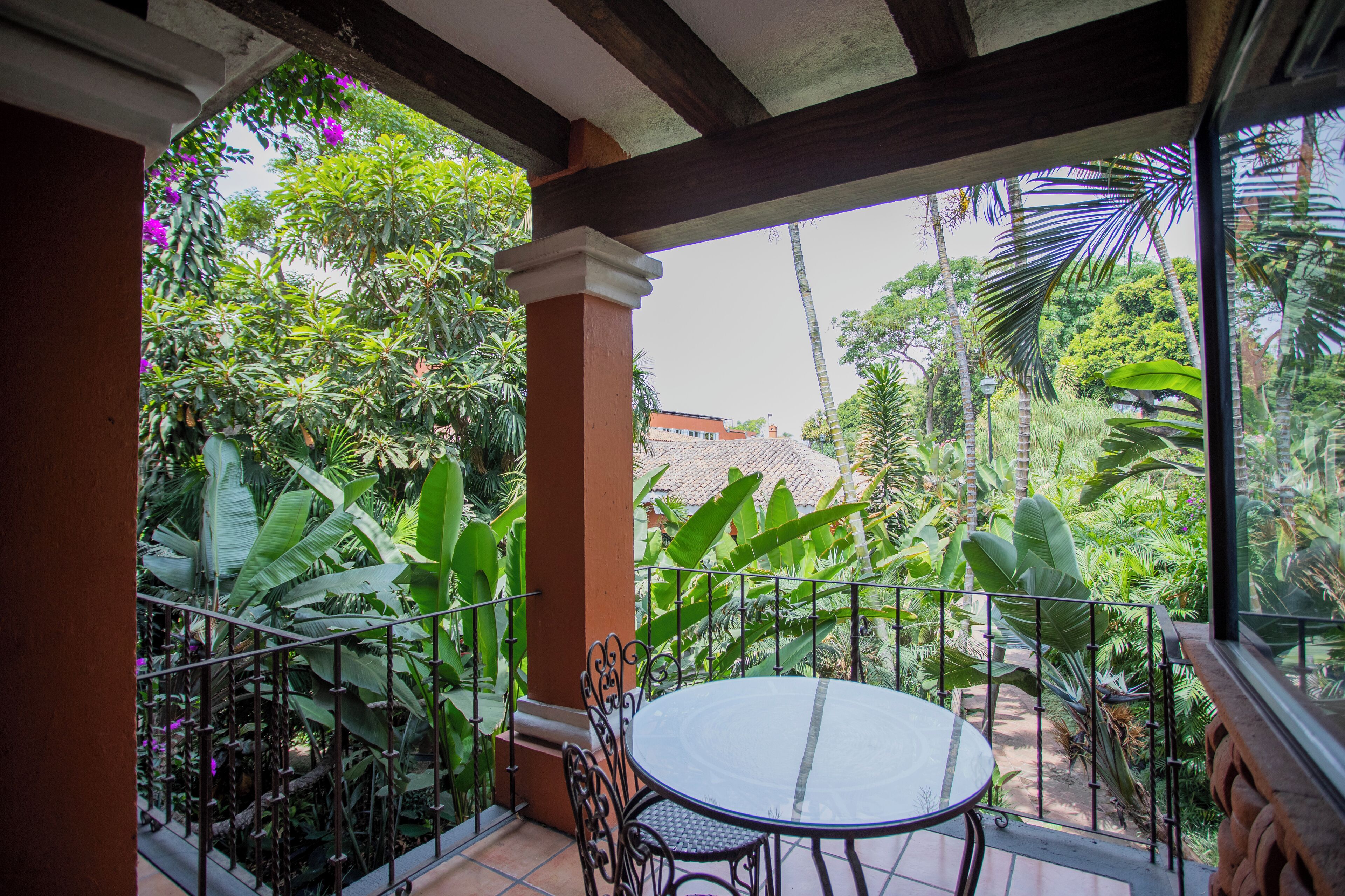 Un balcón con una mesa redonda y sillas, rodeado de exuberante vegetación tropical y una vista de un edificio al fondo.