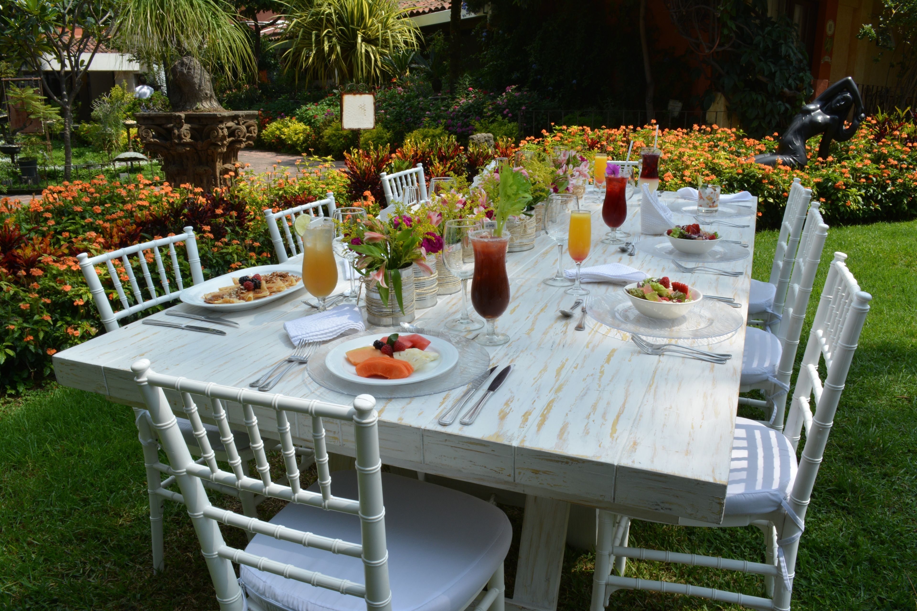 An outdoor dining table set with white chairs, surrounded by a garden with flowers and a statue.